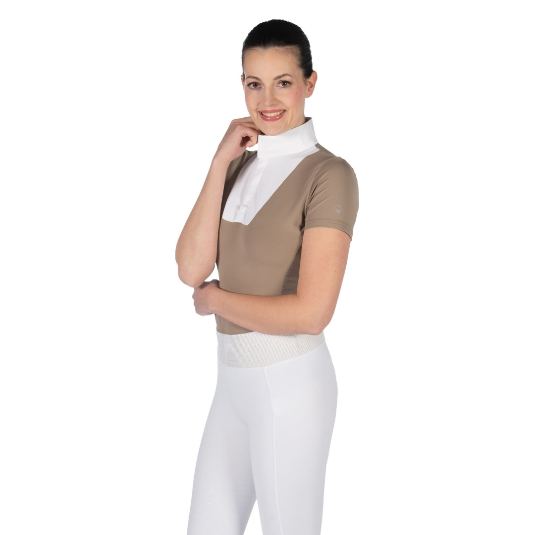 Woman wearing beige equestrian top with a white high-neck collar and white riding pants, posing with her hand near her chin.