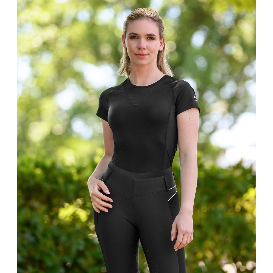 Woman in black athletic top and leggings standing outdoors in a sunlit park with green foliage behind her.