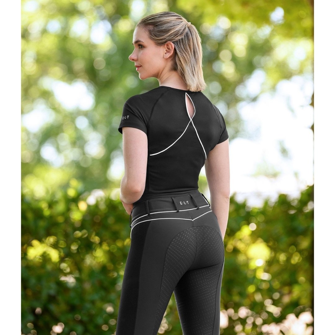 Woman in a black athletic outfit standing outdoors in a sunlit park, back view with white seam lines on the shirt and white piping on high‑waisted leggings.