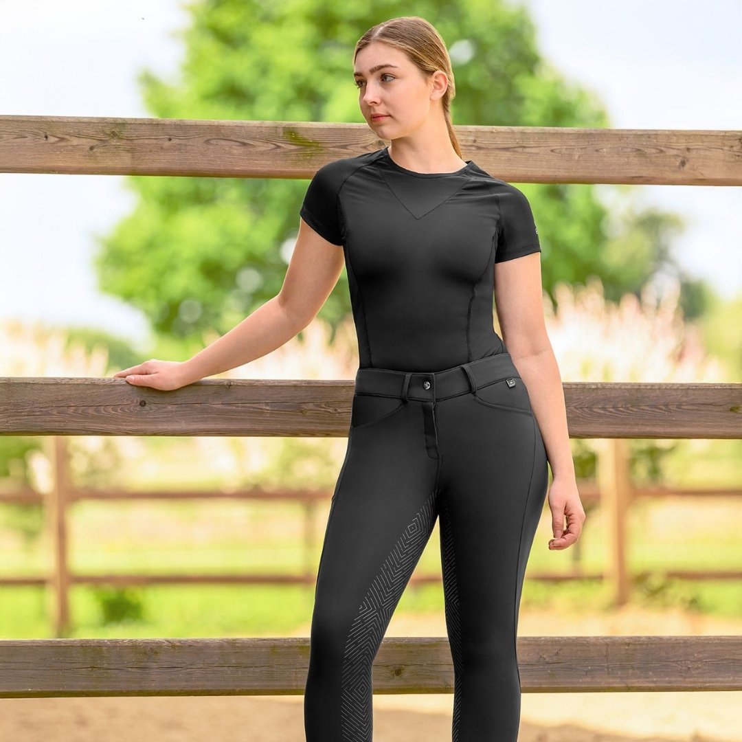 Woman wearing a black athletic shirt and leggings leaning on a wooden fence in a sunny outdoor setting with greenery in the background.