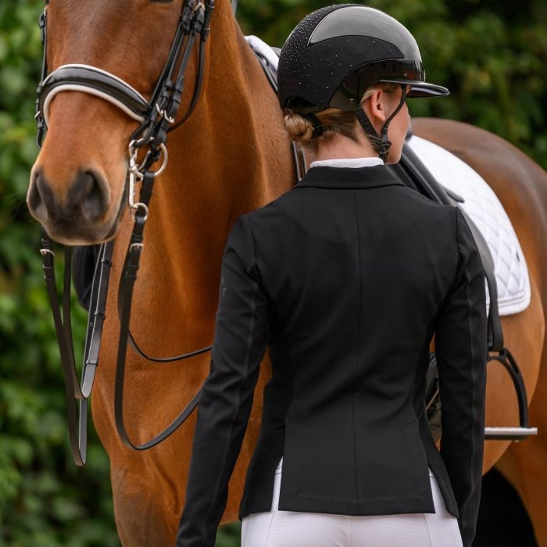 Equestrian in a black riding jacket and white breeches stands beside a brown horse with bridle and saddle, outdoors in greenery.