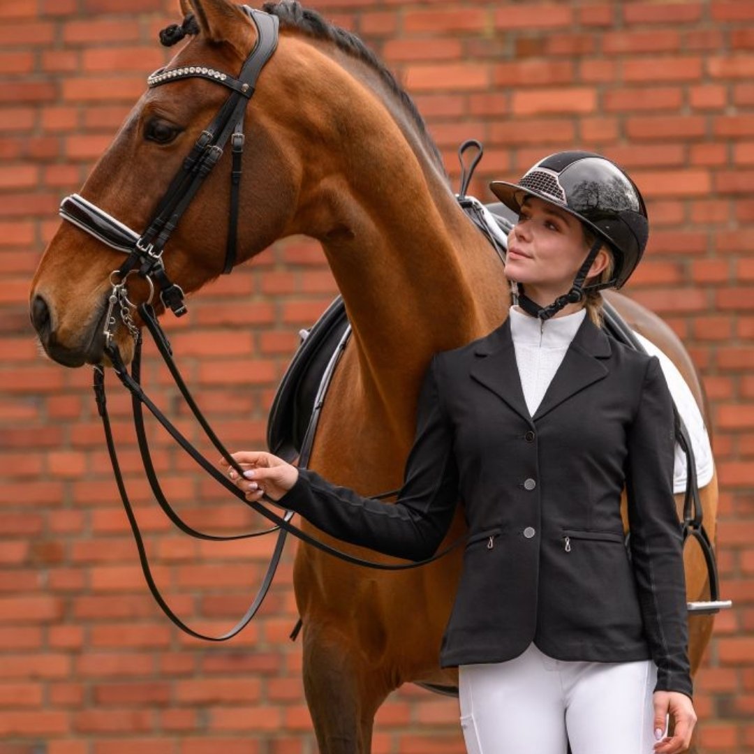Woman in a black riding jacket and helmet stands beside a chestnut horse with reins, against a brick wall behind them.