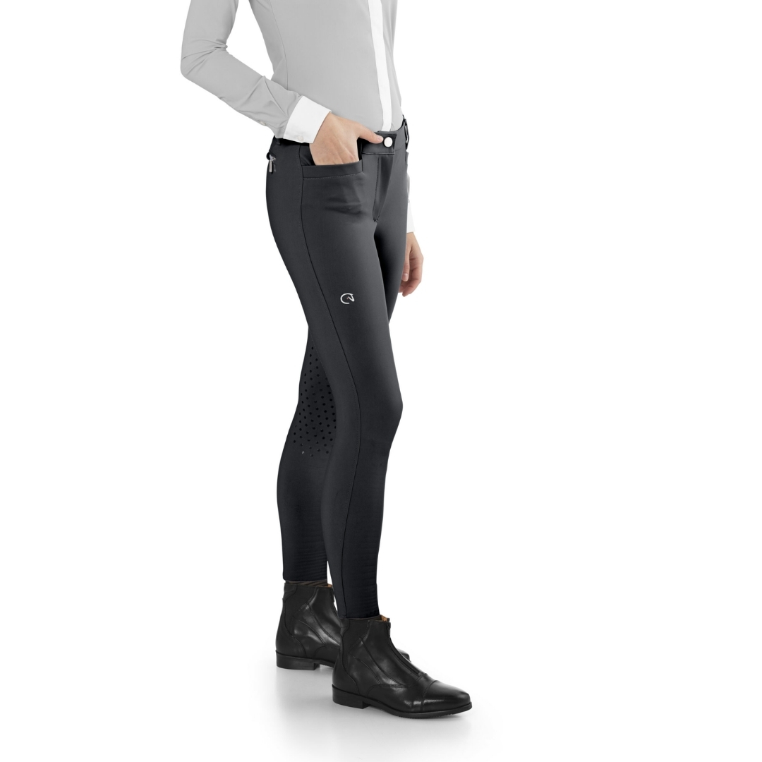 Side view of a person wearing black riding breeches with perforated panels, white shirt, and black ankle boots, showing a small logo on the thigh.