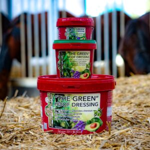 Stacked red containers of 'The Green Hoof Dressing' in a stable with horses in the background; straw on the floor and green lids visible on top container.