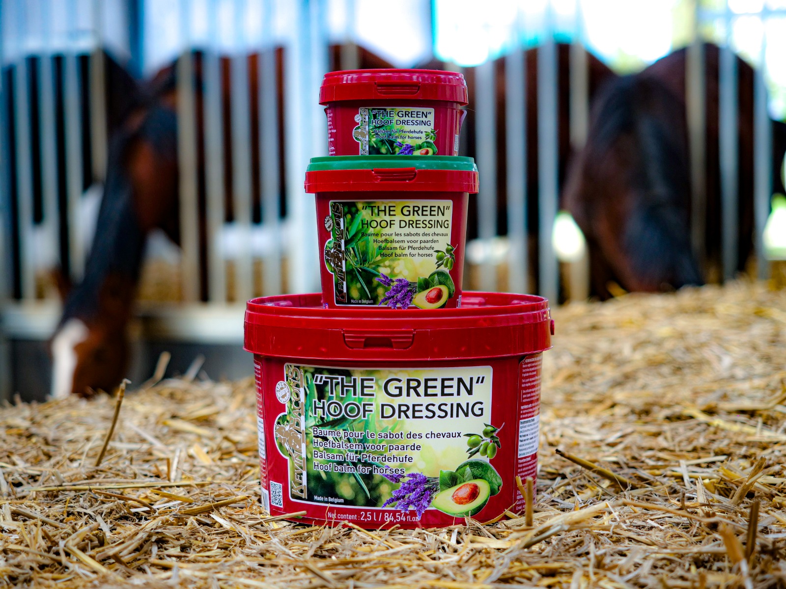 Stacked red containers of 'The Green Hoof Dressing' in a stable with horses in the background; straw on the floor and green lids visible on top container.