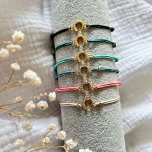 Six colorful cord bracelets with gold horseshoe charms encrusted with crystals, stacked on a gray display roll (black, green, teal, blue, pink, cream).