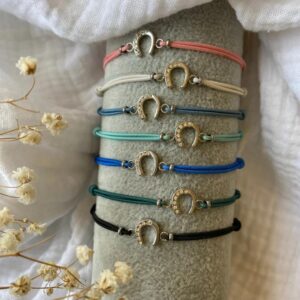Shelf of colorful cord bracelets, each with a gold horseshoe charm adorned with small crystals, arranged on a gray display.
