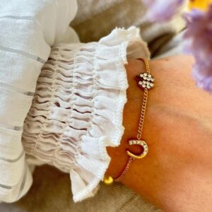 Gold chain bracelet with a sparkling diamond-encrusted crescent charm on a wrist, beside a white frilled sleeve.