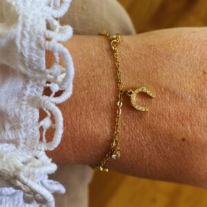 Gold bracelet with a jewel-encrusted crescent moon charm on a wrist with white lace sleeve.